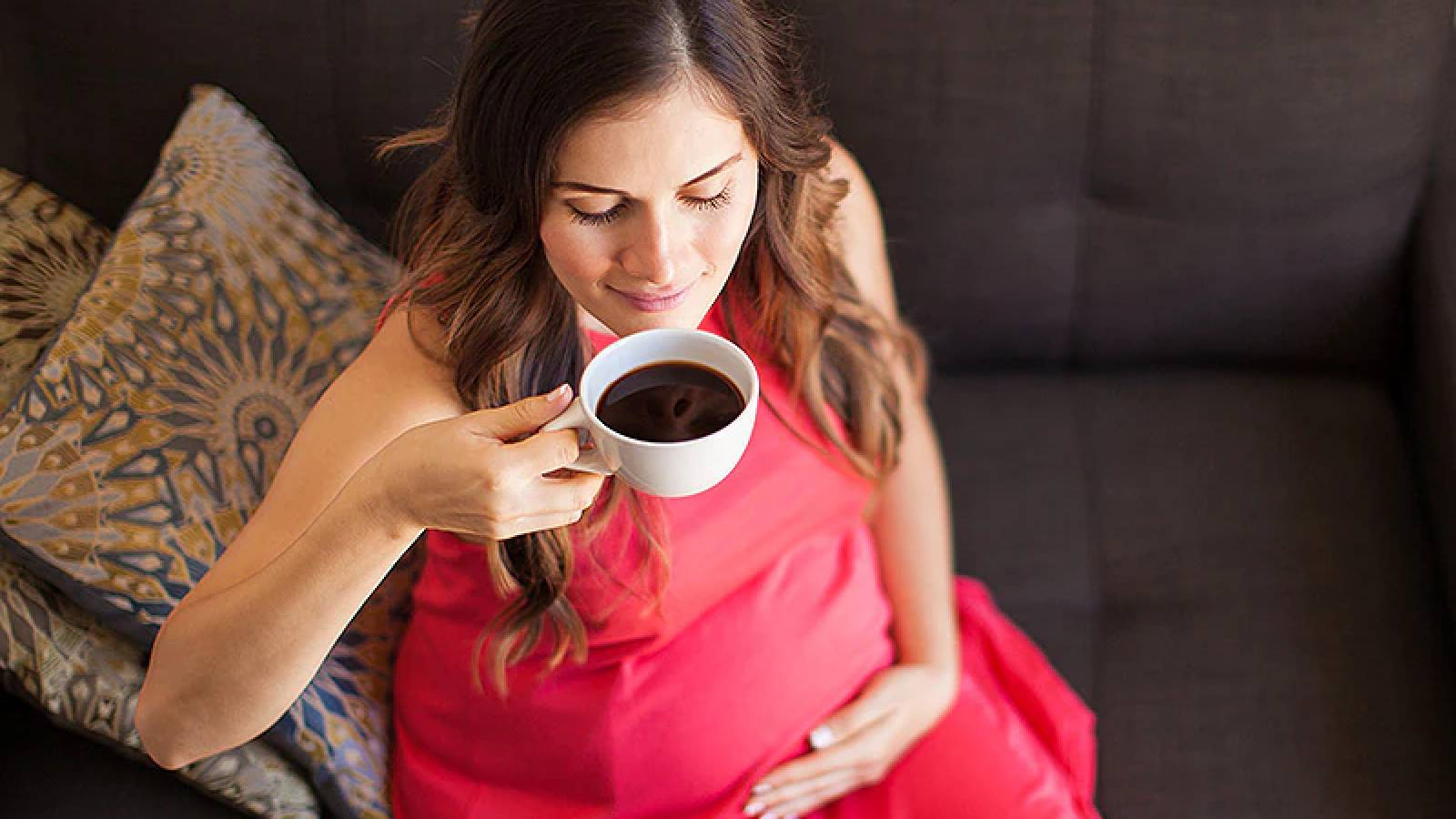 امرأة حامل ترتدي فستان احمر تجلس و تحمل بيدها فنجان من القهزة وتقربه من فمها لتشربه ويدها اليسرى تضعها على بطنها 