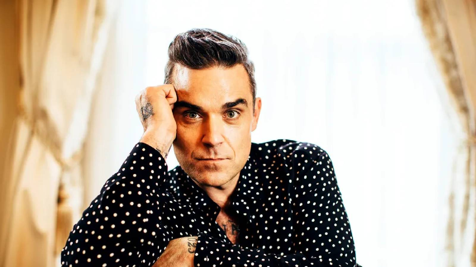 Robbie Williams يرتدي قميص اسود منقط بالابيض وهو متكئ على الكرس وييسند رأسه بيده وخلفه نافذة وعليها ستائر 