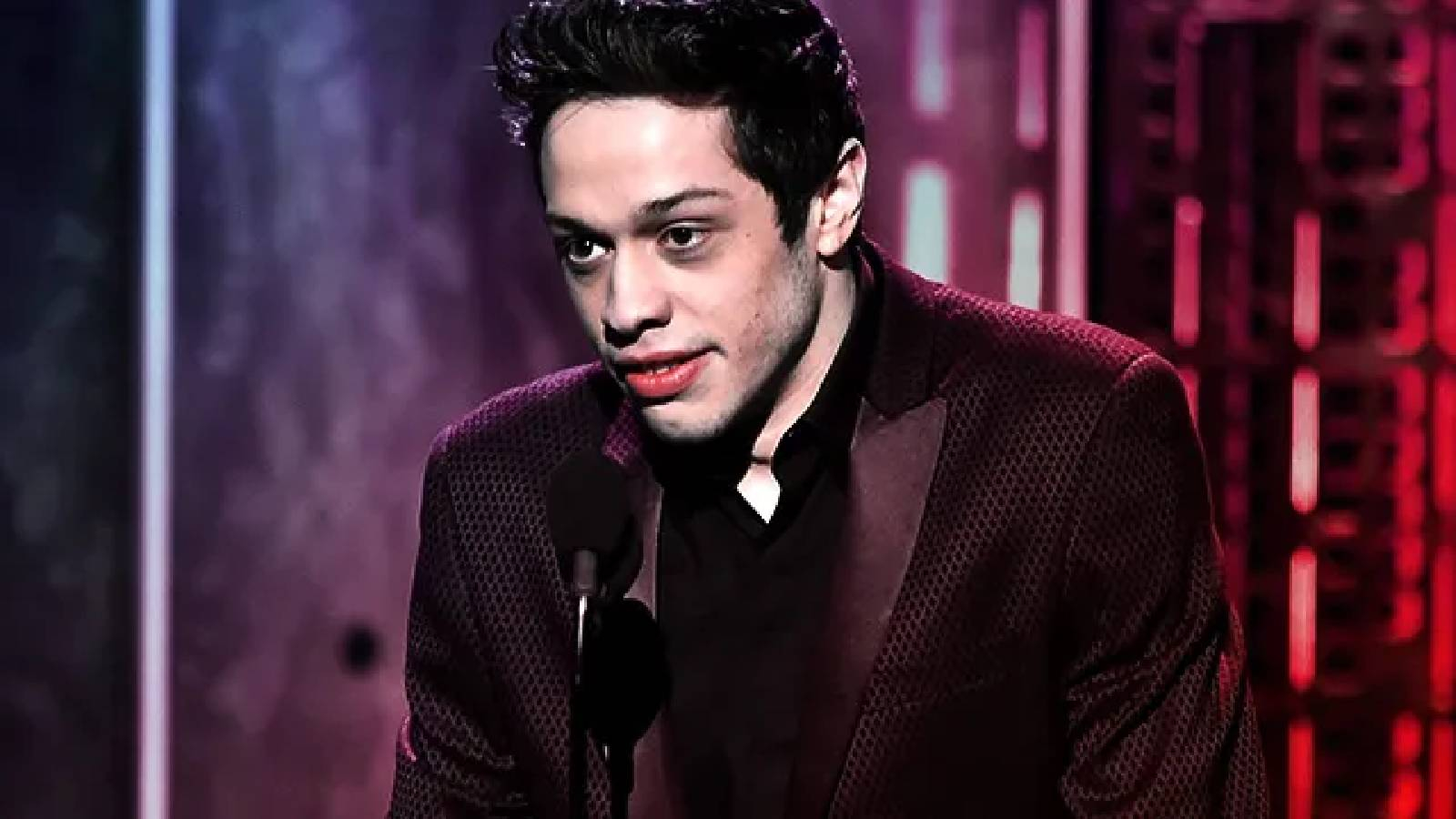 Pete Davidson وهو يرتدي بدلة سوداء ويقف امام الميكروفون وخلفه ديكور مسلطة عليه أضواء حمراء 