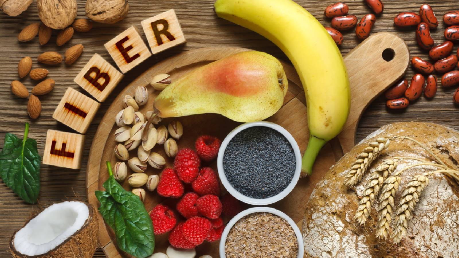 خمس مكعبات كل مكعب مكتوب عليه حرف موضوعين على طاولة لتشكيل كلمة FIBER وبجانبهم طبق خشب موضوع عليه الفستق الحلبي و الفريز و بدور الشيا والسبانخ و حوله الجوز وجوز الهند والقمح و غيرها 