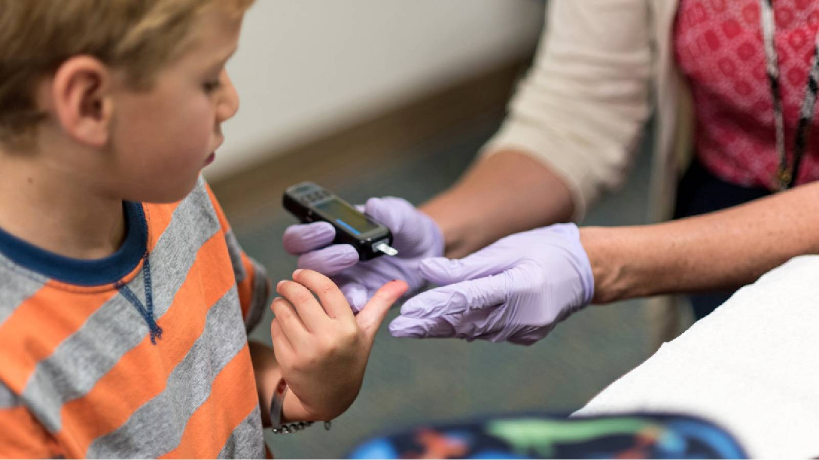 طفل يرتدي تيشيرت مخططة يمد ابهامه للمرضة التي ترتدي قفازات طبية و تحمل بيدها جهاز فحص السكر 