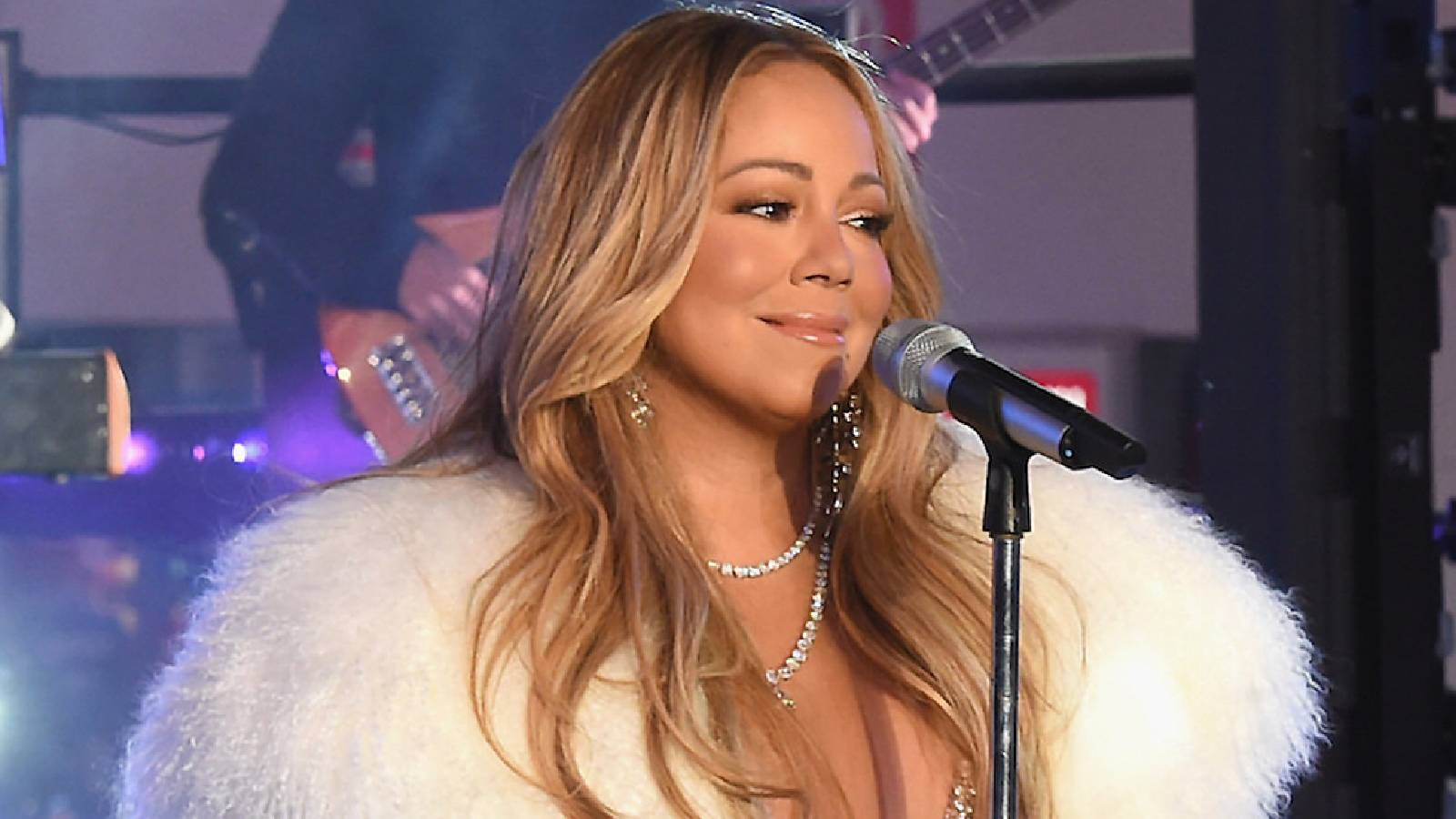 Mariah Carey تقف على المسرح أما الميكروفون وخلفها عازف الغيتار وهي ترتدي جاكيت من الفرو الابيض مسدلة شعرها ع