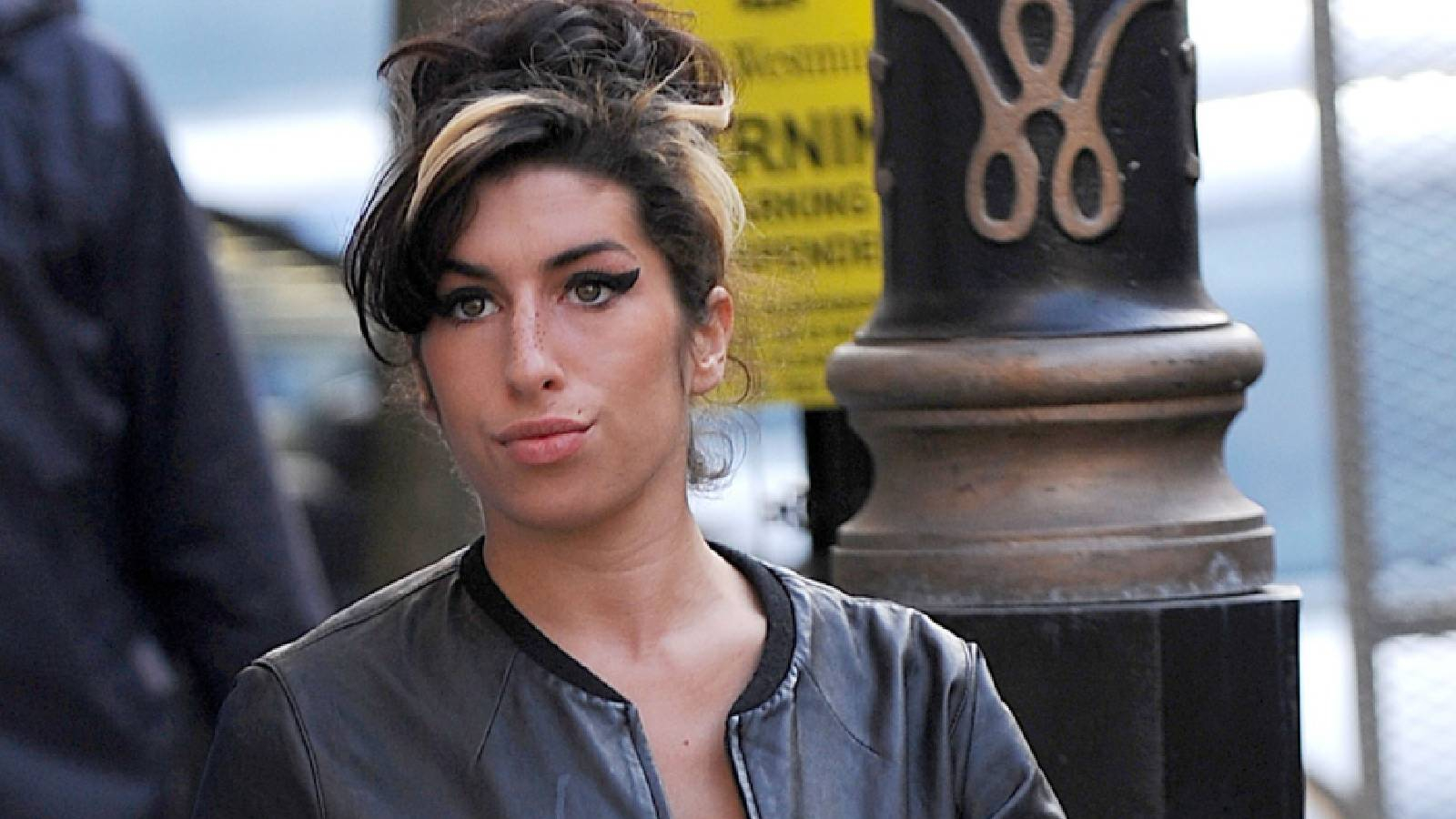 Amy Winehouse تمشي في الشارع مرتدية سترة سوداء وترفع شعرها وخلفها عمود اسود ولوحة إعلان صفراء 