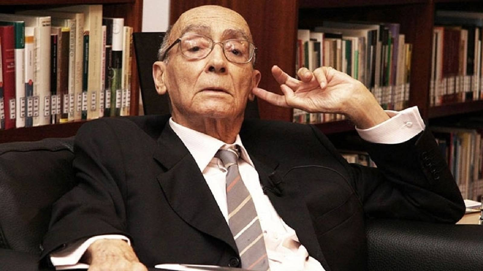 الكاتب جوزيه ساراماغو وهو جالس في مكتبه رافعا يده الى راسه وخلفه مكتبته المليئة بالكتب 