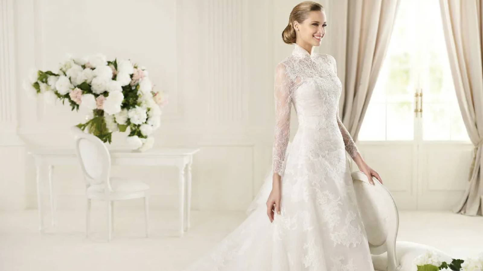 في مكان انيق باللون الابيض وفيه ورود تقف عروس ترتدي فستان ابيض انيق بأكام شيفون و هي مستندة الى كرسي ابيض 