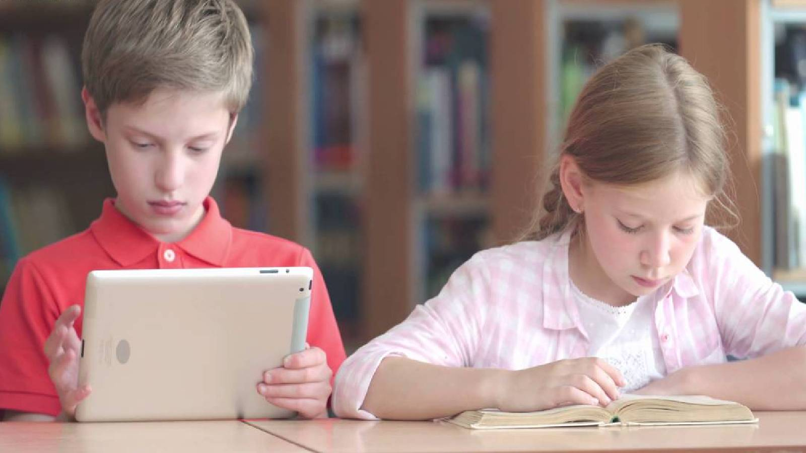 طفلين يجلسان الى طاولة الطفلة الاولى تقرأ بكتاب ورقي و الطفل الثاني يحمل تاب و يقرا قراءة الكترونية 